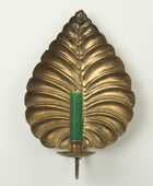 A vintage-style brass leaf-shaped wall sconce holder with a green candle inserted in the center, set against a plain off-white background.