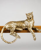 A metallic leopard figurine with a textured, speckled gold finish, resting on a wooden bar against a neutral gray background.