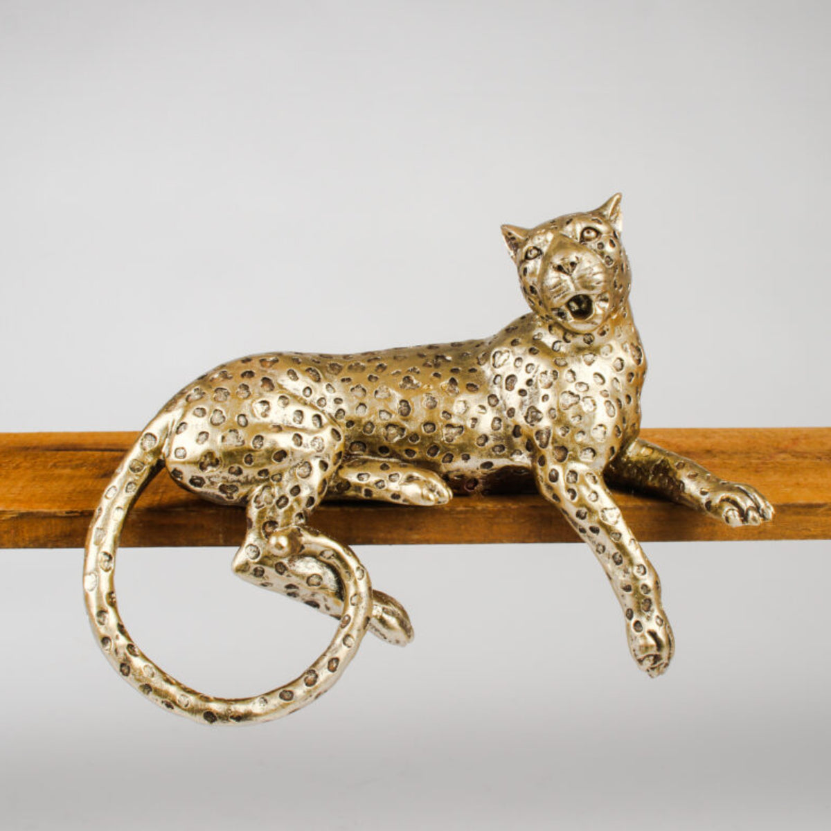 A metallic leopard figurine with a textured, speckled gold finish, resting on a wooden bar against a neutral gray background.