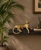 A metallic gold leopard figurine resting on a dark wooden floating shelf in a styled interior setting with a potted palm plant, a framed print, and decorative books.