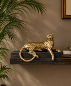 A metallic gold leopard figurine is displayed on a dark wooden floating shelf in a styled interior setting. A potted palm plant is visible on the left, and a book and diffuser sit on the right against a warm beige wall.