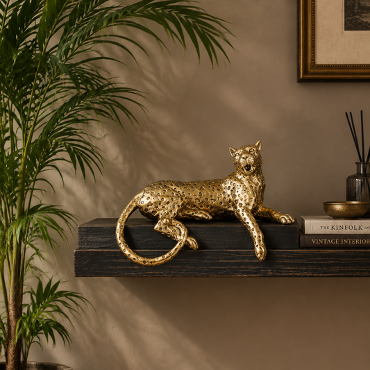 A metallic gold leopard figurine is displayed on a dark wooden floating shelf in a styled interior setting. A potted palm plant is visible on the left, and a book and diffuser sit on the right against a warm beige wall.