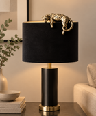 A modern table lamp with a black cylindrical base and a black fabric drum shade, topped with a gold-finished metal rod holding a gold leopard figurine. The lamp sits on a dark side table in a warm, contemporary living room setting.