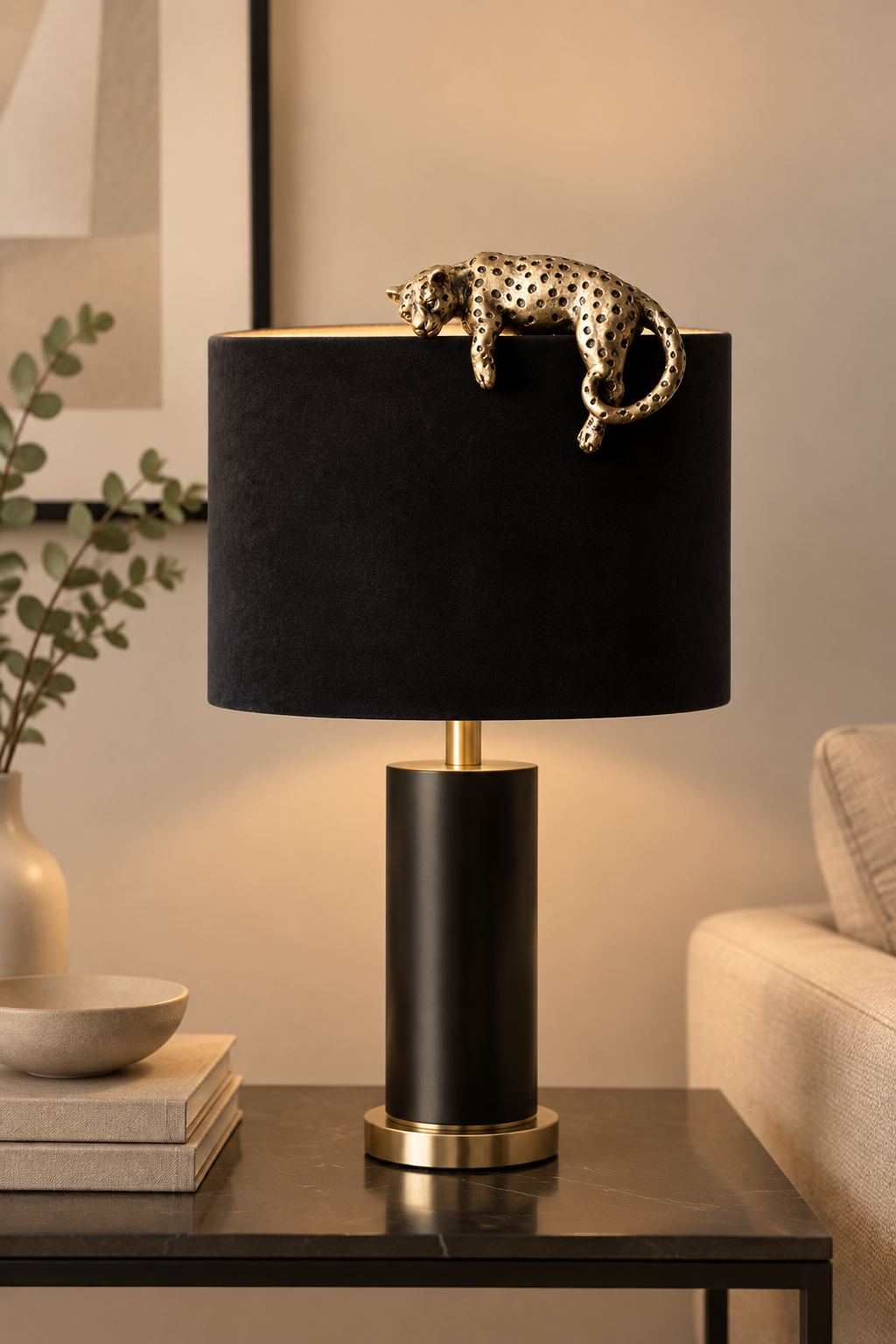 A modern table lamp with a black cylindrical base and a black fabric drum shade, topped with a gold-finished metal rod holding a gold leopard figurine. The lamp sits on a dark side table in a warm, contemporary living room setting.