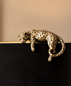 A metallic leopard figurine with a perforated, textured finish in a gold/bronze tone, resting across the top of a black cylindrical lamp base against a warm beige background.