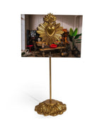 A gold-colored freestanding picture frame with an ornate sculpted base and a central stem, holding a rectangular photograph of a decorative interior scene with a large gold sacred heart motif.