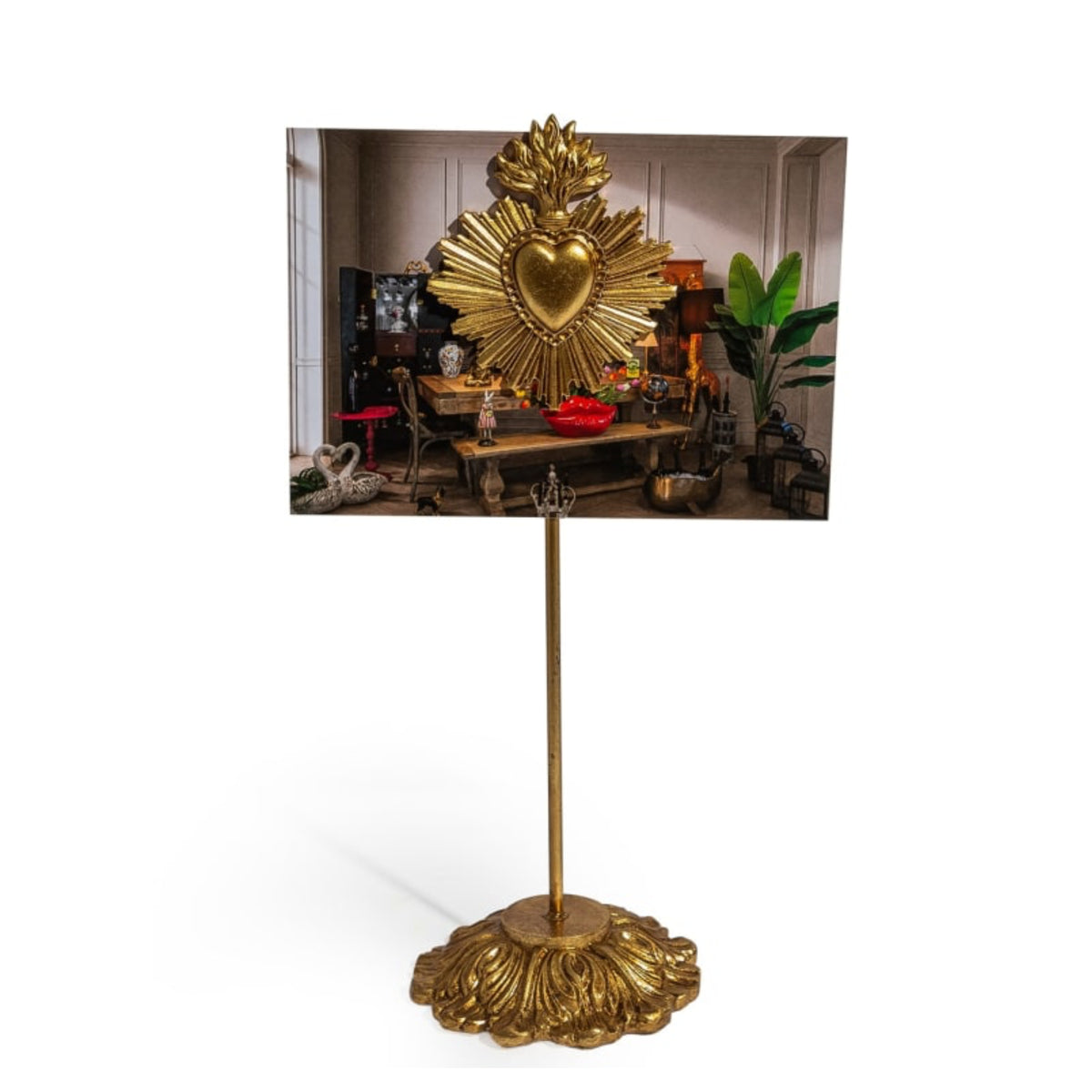 A gold-colored freestanding picture frame with an ornate sculpted base and a central stem, holding a rectangular photograph of a decorative interior scene with a large gold sacred heart motif.
