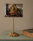A brass Sacred Heart of Jesus tabletop display stand holding a framed art print, styled on a distressed teal wooden table next to an antique book against a textured beige wall.