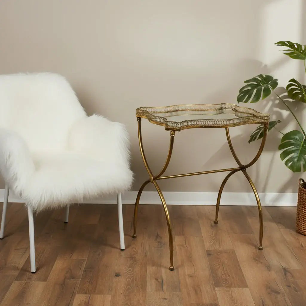 Gold side table with glass top next to a white fur chair on a wooden floor.