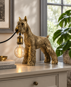 A decorative dog-shaped table lamp in a metallic gold finish stands on a console table in a styled room. The lamp is shaped like a terrier dog and holds a glowing light bulb in its mouth, with a black cord extending to the left.