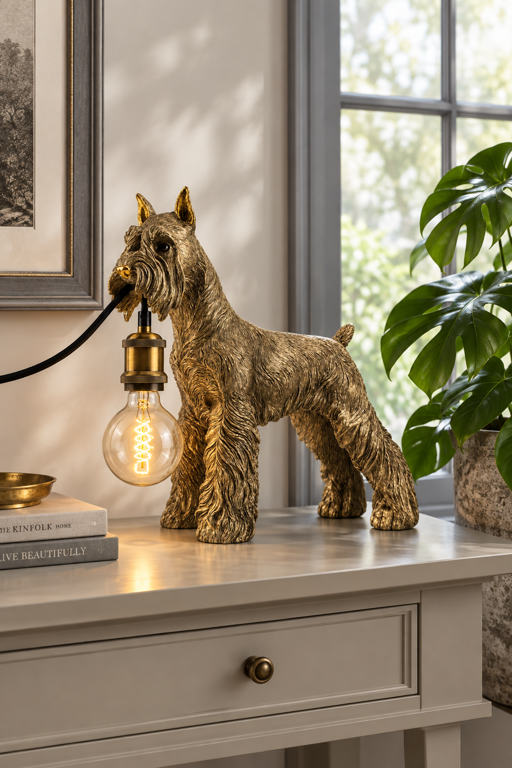 A decorative dog-shaped table lamp in a metallic gold finish stands on a console table in a styled room. The lamp is shaped like a terrier dog and holds a glowing light bulb in its mouth, with a black cord extending to the left.