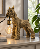 A decorative dog-shaped table lamp in a metallic gold finish stands on a light-colored cabinet beside a potted plant. The lamp has a clear bulb with a visible filament and a black cord extending out of the dog's mouth.