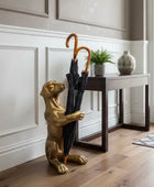 Gold dog-shaped umbrella stand holding a black umbrella in a room with wooden flooring and a small table.