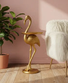 Gold flamingo side table next to a white furry chair and potted plant against a pink wall.