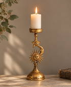 A decorative gold-toned metal candlestick holder with sculpted celestial motifs, including a sun, moon, and stars, holding a lit white pillar candle. The holder is styled on a wooden surface with a neutral background, a potted plant on the left, and an old book on the right.