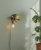 Gold turtle-shaped wall lamp with a lit bulb on a light gray wall, next to a potted plant.
