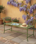 Green metal bench with a hat and book in a garden setting with wisteria and potted plants.