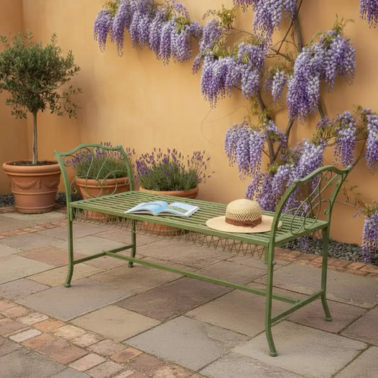 Green metal bench with a hat and book in a garden setting with wisteria and potted plants.