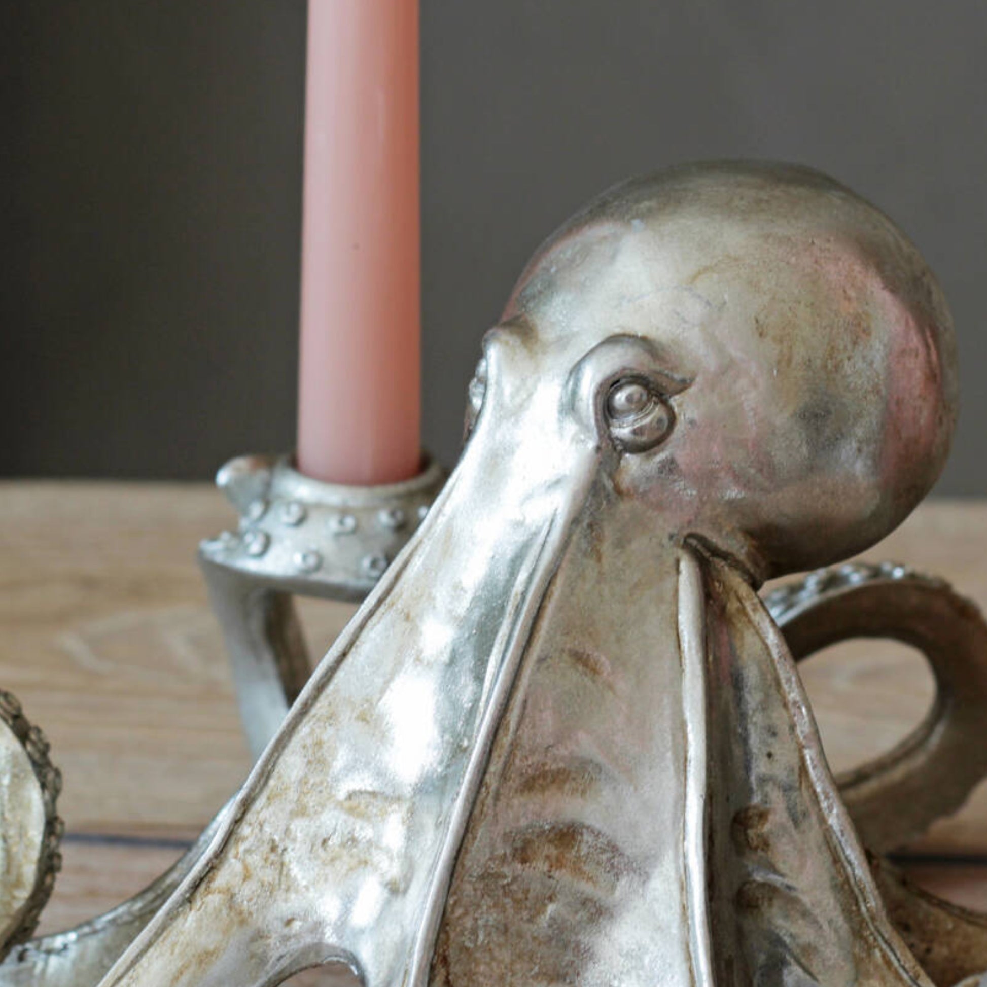 A close-up of a metallic octopus-shaped candle holder with a pink candle inserted, set on a wooden surface against a dark gray background.