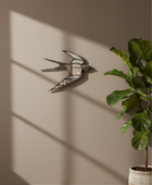 A wall-mounted bird-shaped mirror with a metallic frame and glass panels, displayed on a beige wall with dramatic window shadows. A potted green plant stands to the right, and the scene is styled in a modern interior setting.