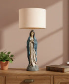 Table lamp with a figurine on a wooden surface next to a plant and book.