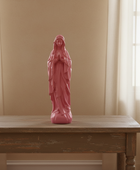 A pink religious figurine of the Virgin Mary standing on a rustic wooden table against a beige wall with sheer curtains.