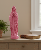 A pink religious figurine of the Virgin Mary standing on a light wood console table, with a potted fern on the left and a vintage book on the right, set against a beige wall and a window.