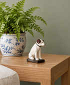 A small white and brown dog figurine sits on a light wood table next to a potted fern in a blue-and-white ceramic pot against a muted green wall.