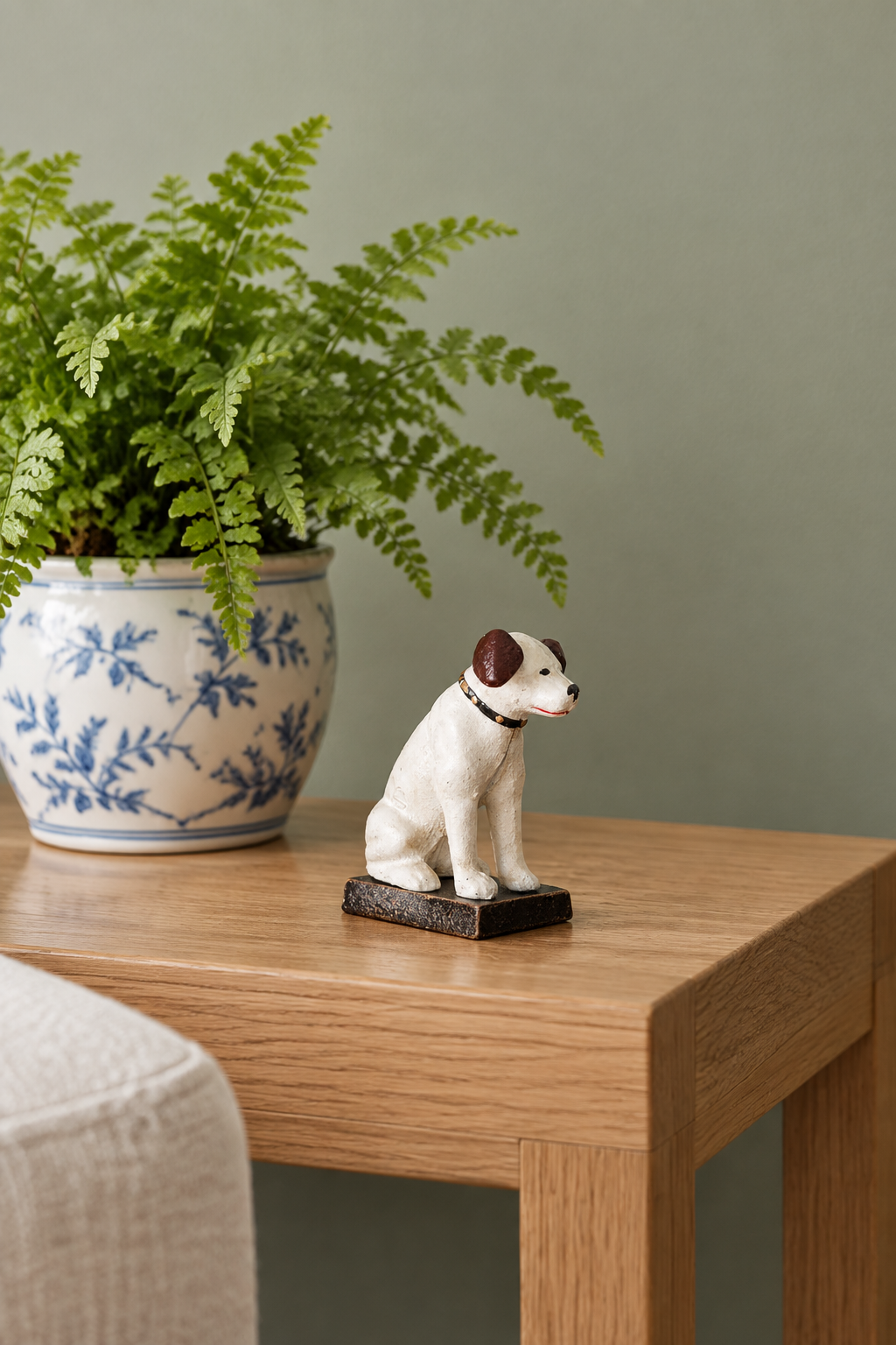 A small white and brown dog figurine sits on a light wood table next to a potted fern in a blue-and-white ceramic pot against a muted green wall.