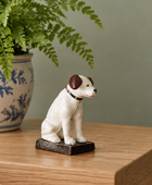 A small ceramic dog figurine in white with brown ears sits on a dark rectangular base on a wooden table, with a potted fern in the background.