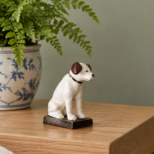 A small ceramic dog figurine in white with brown ears sits on a dark rectangular base on a wooden table, with a potted fern in the background.