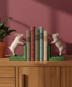 Pig-shaped bookends on a wooden shelf with books and a plant against a pink wall.