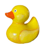 A glossy yellow rubber duck with an orange beak, photographed on a cardboard surface with storage boxes and shelving in the background.