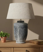 Decorative lamp with textured base and beige shade in a room setting with framed artwork and mugs.