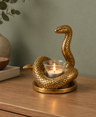 A gold-toned snake-shaped candle holder with a lit tealight candle in a clear glass holder, displayed on a wooden table against a muted green wall.