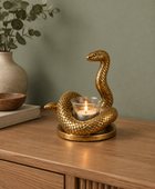 A gold-toned snake figurine candle holder with a lit tealight candle in a glass holder, displayed on a wooden sideboard against a sage green wall.