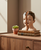 Two mannequin heads on books with a potted plant on a wooden surface near a window.