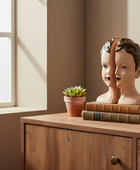 Two mannequin heads on books with a potted plant on a wooden surface near a window.
