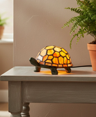 A turtle-shaped stained glass lamp glowing with warm yellow and orange light, placed on a gray wooden table next to a potted fern in a real indoor setting.