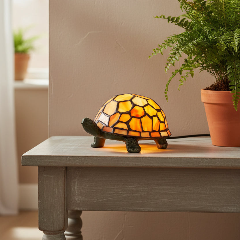 A turtle-shaped stained glass lamp glowing with warm yellow and orange light, placed on a gray wooden table next to a potted fern in a real indoor setting.