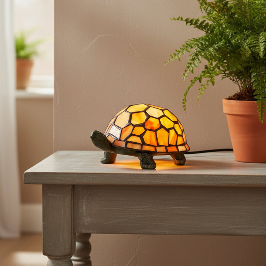 A turtle-shaped stained glass lamp glowing with warm yellow and orange light, placed on a gray wooden table next to a potted fern in a real indoor setting.
