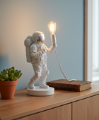 Astronaut lamp holding a light bulb on a wooden surface with a plant in the background.