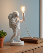 Spaceman lamp with a light bulb on a wooden surface next to a plant and book