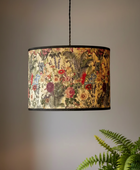 A cylindrical pendant lampshade with a dense botanical and floral print in red, green, blue, and beige, hanging against a neutral gray wall with a green fern in the foreground.
