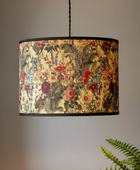 A drum pendant light with a vintage botanical patterned fabric shade, hanging against a neutral gray wall with a green fern in the foreground.