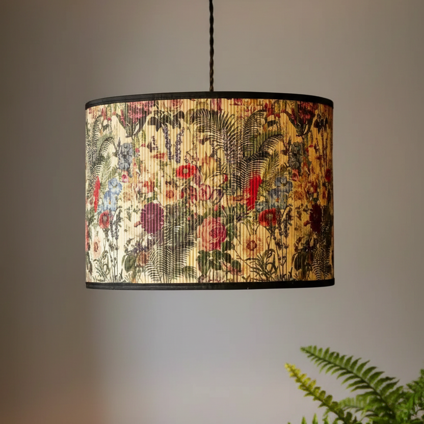A drum pendant light with a vintage botanical patterned fabric shade, hanging against a neutral gray wall with a green fern in the foreground.