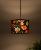 A hanging drum pendant lamp with a dark background and a colorful floral print, shown lit against a warm beige wall with a green fern in the lower right corner.