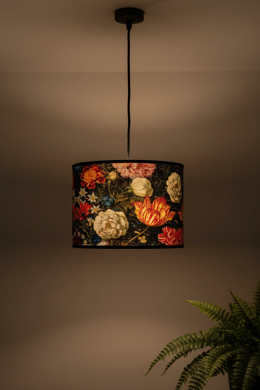 A hanging drum pendant lamp with a dark background and a colorful floral print, shown lit against a warm beige wall with a green fern in the lower right corner.