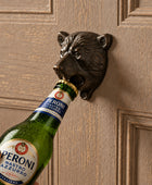 A black bear head bottle opener mounted on a beige paneled door, being used to open a gold foil-wrapped bottle. A hand is visible holding the bottle.