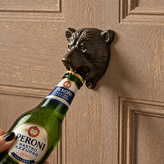 A black bear head bottle opener mounted on a beige paneled door, being used to open a gold foil-wrapped bottle. A hand is visible holding the bottle.